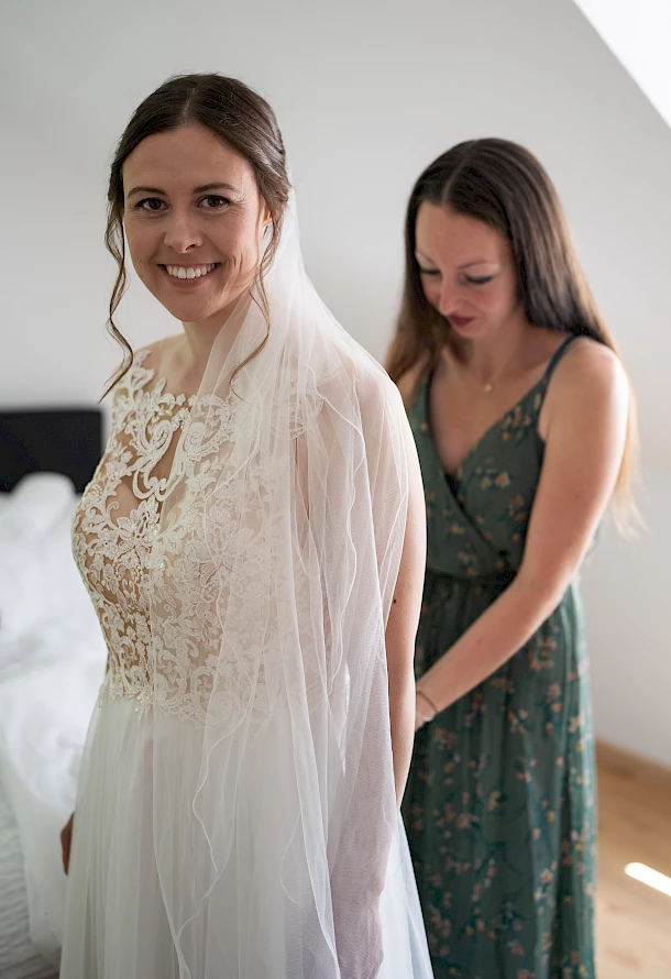 Braut wird von Trauzeugin beim Kleid anziehen unterstützt